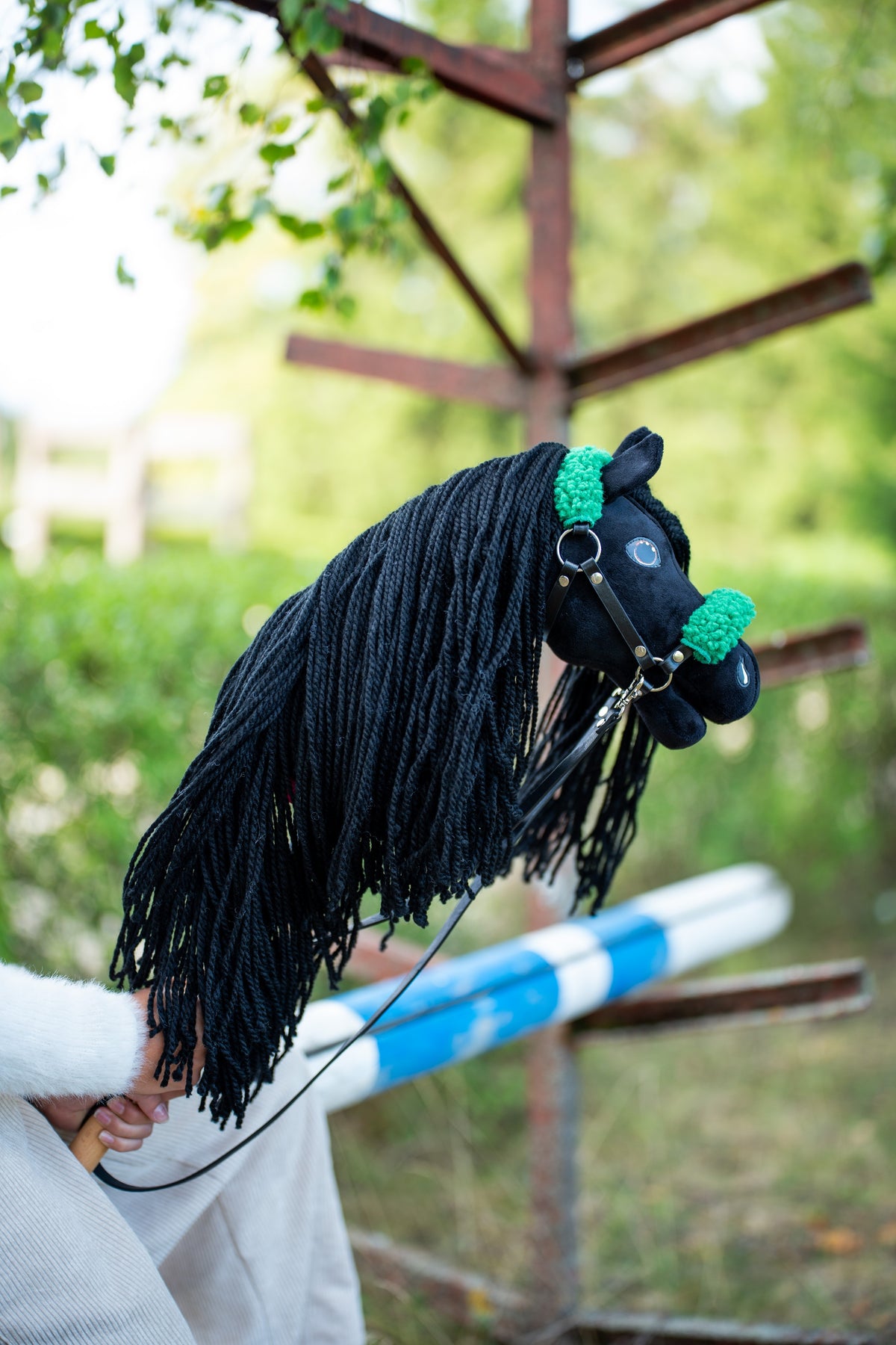 Hlava a dlouhá hříva Hobby Horse Maximus s černou ohlávkou od LarDen jsou zvednuté u dřevěné překážky, v pozadí je zeleň, s černým plyšem a černou přízovou hřívou, s černou koženkovou ohlávkou se zeleným kožíškem a černými koženkovými otěžemi