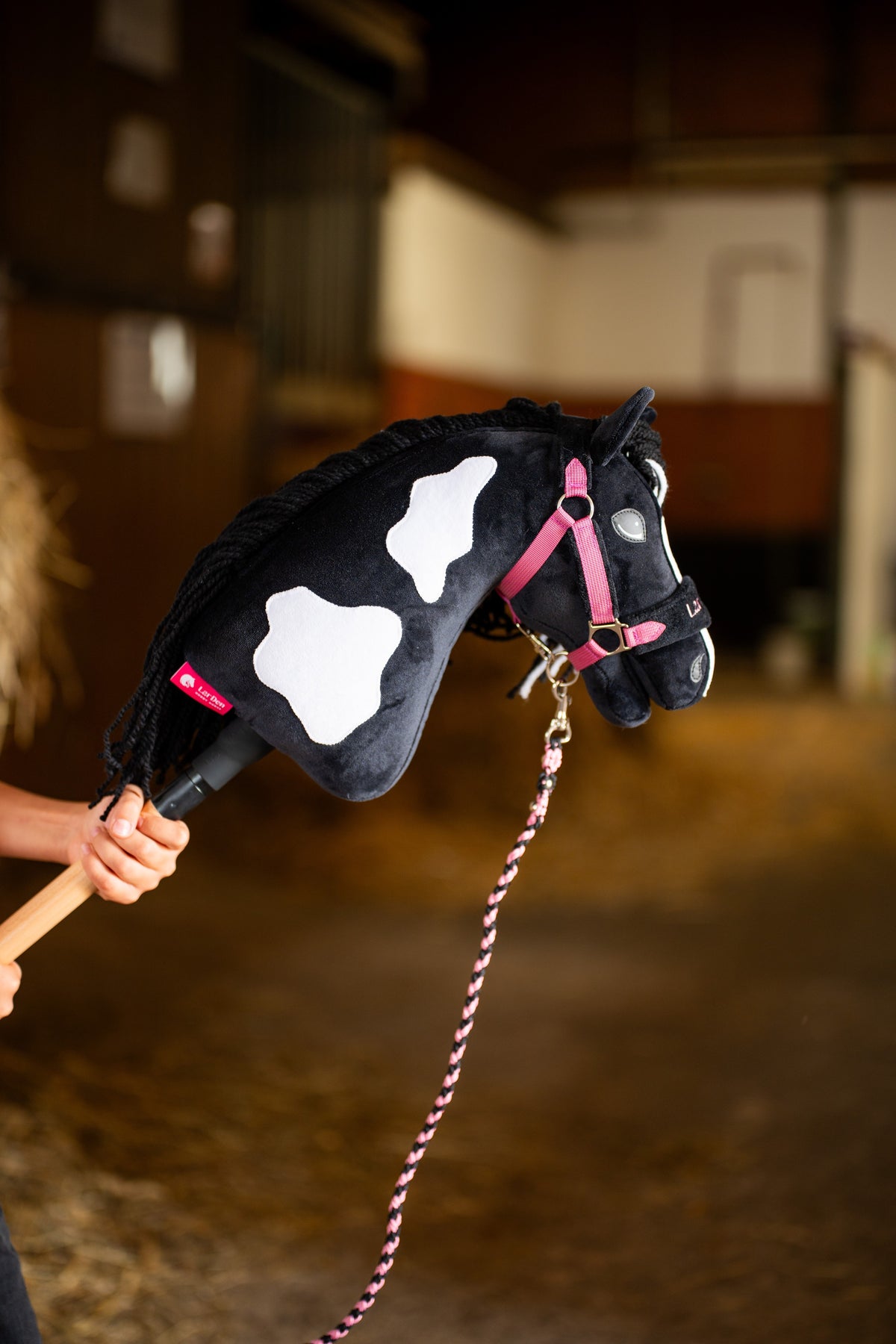Detail photo: the head and neck of the Hobby Horse Merlin with Black Halter by Hobby Horse LarDen held up inside a stable - black plush with white patches, black yarn mane - shown here with the LarDen Exclusive Pink set and its braided pink lead rope.