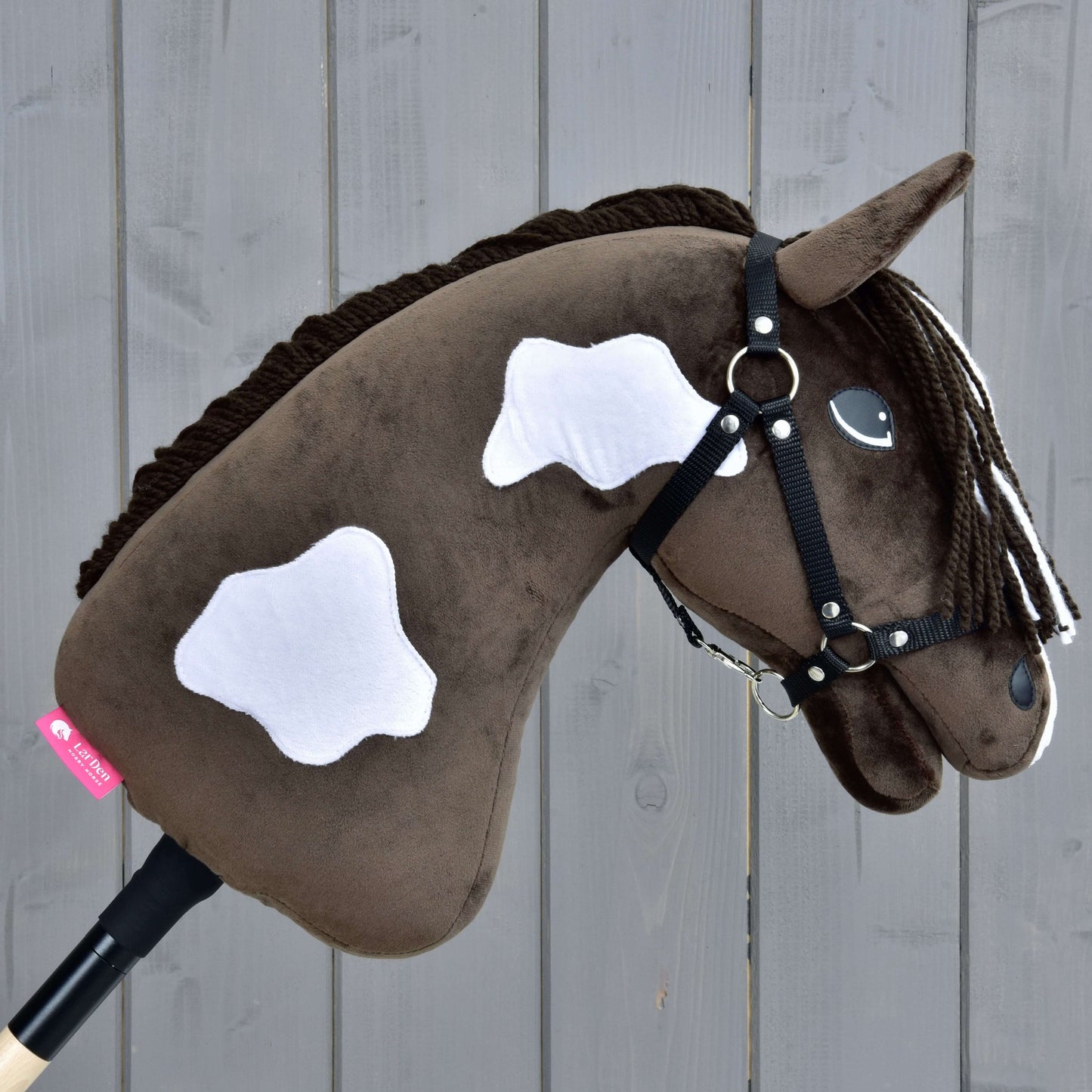 Hobby Horse Monte s černou ohlávkou od Hobby Horse LarDen je hnědý plyšový Hobby Horse s bílými skvrnami a černou ohlávkou, upevněný na dřevěné tyči, zobrazený před šedým pozadím z dřevěných prken.