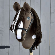 Ručně vyráběný Hobby Horse Monte s černou ohlávkou od Hobby Horse LarDen má hnědobílou srst inspirovanou American Paint Horse, černou ohlávku, dlouhou přízovou hřívu a je upevněný na dřevěné tyči před šedým dřevěným pozadím.