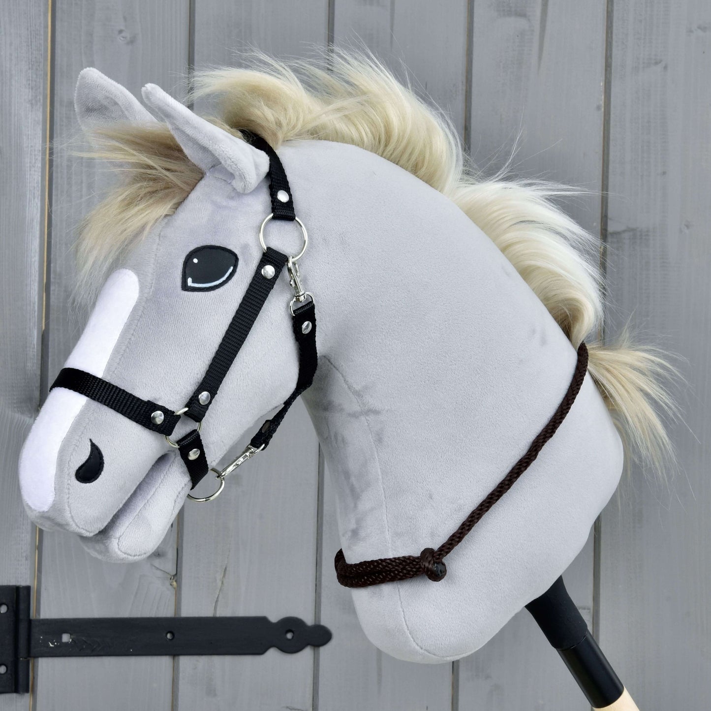 Nákrčník Chocolate od Hobby Horse LarDen se šedým plyšovým Hobby Horse, černou uzdečkou a krémovou provazovou hřívou na dřevěné tyči, na pozadí šedého dřevěného panelu.