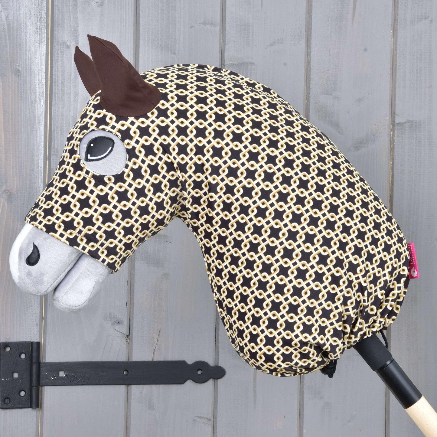 Hobby Horse ochranný oblek Luxus Design od Hobby Horse LarDen s černobéžovou vzorovanou hlavou, plstěnýma ušima, našitým okem, bílým čenichem a pratelnými materiály, nasazený na dřevěné tyči před šedou stěnou.