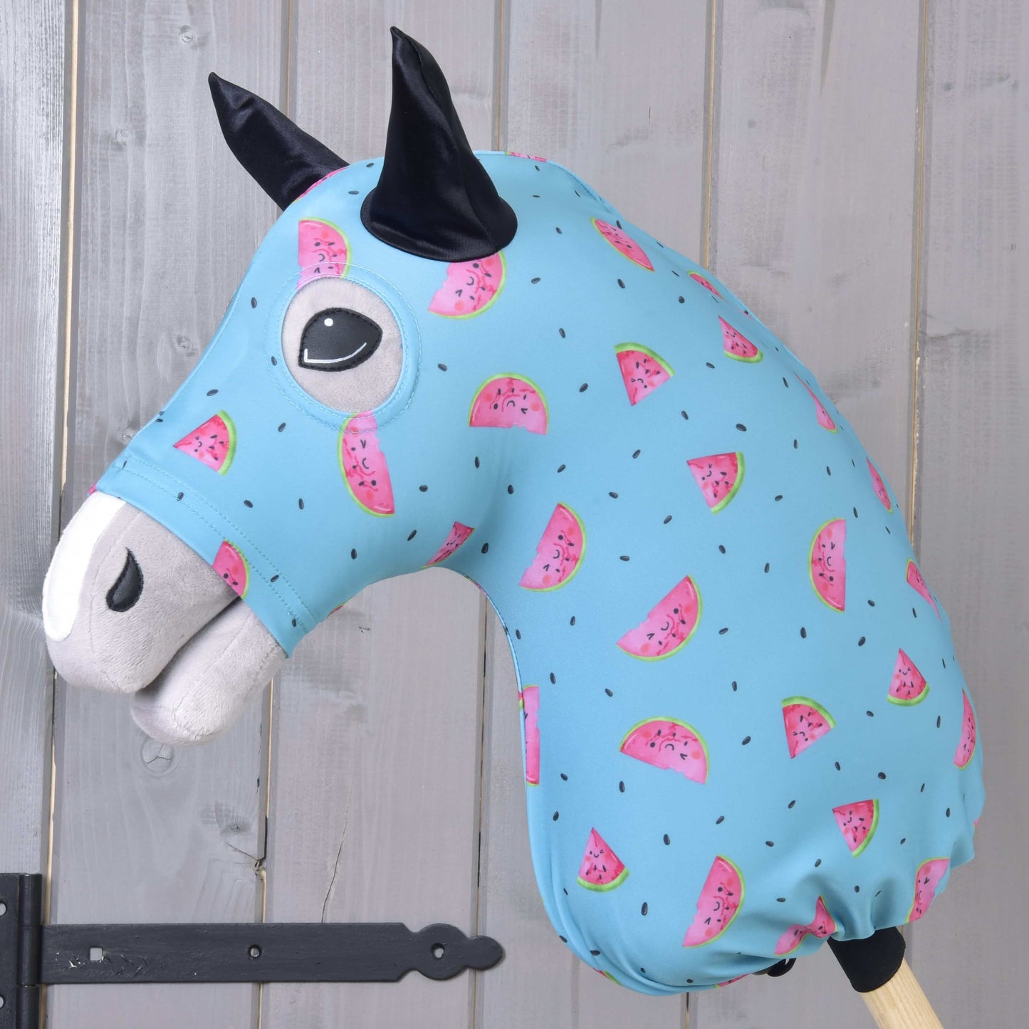 Hobby Horse ochranný oblek Melon (Lycra) od Hobby Horse LarDen má pratelný modrý potah s růžovými plátky melounu a černými semínky, navržený pro plyšové hlavy Hobby Horse. Ideální pro ochranu, zobrazený na dřevěné tyči před světlou stěnou z dřevěných panelů.