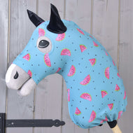 Hobby Horse ochranný oblek Melon (Lycra) od Hobby Horse LarDen má pratelný modrý potah s růžovými plátky melounu a černými semínky, navržený pro plyšové hlavy Hobby Horse. Ideální pro ochranu, zobrazený na dřevěné tyči před světlou stěnou z dřevěných panelů.