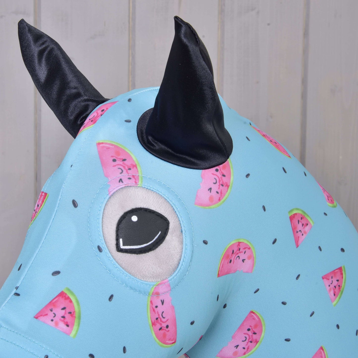 Detailní záběr na Hobby Horse ochranný oblek Melon (Lycra) od Hobby Horse LarDen, světle modrý pratelný lycrový potah se vzorem melounů, jedním velkým vyšívaným okem a dvěma černými látkovými ušima, na světlém dřevěném pozadí.