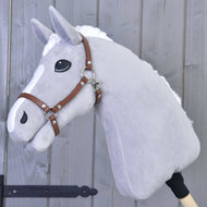 Šedý plyšový Hobby Horse LarDen s bílou hřívou a odznaky na nose má hnědou koženkovou ohlávku, je nasazený na dřevěné tyči a zobrazený před šedou dřevěnou stěnou.