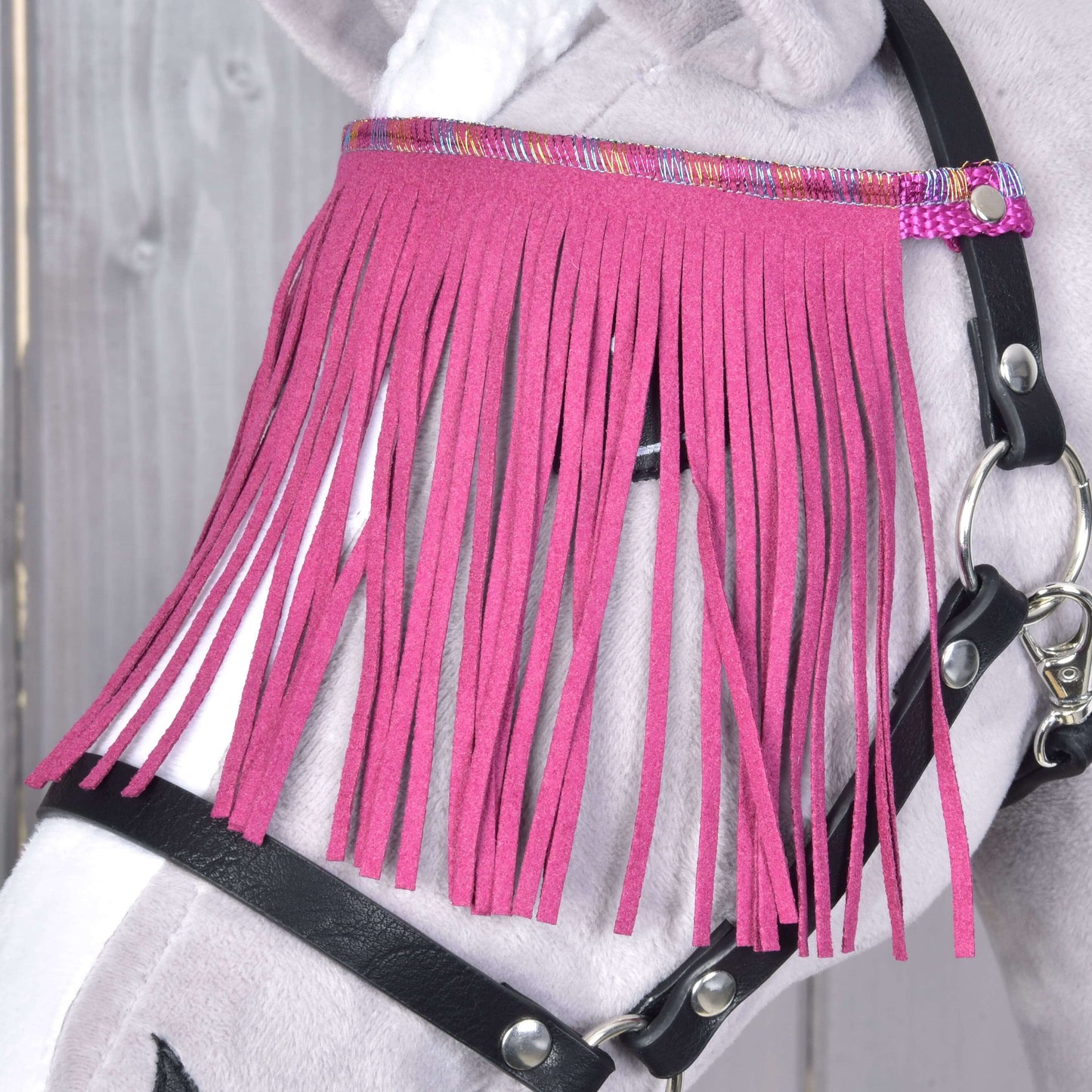 Detail Hobby Horse LarDen „Ohlávka černá s pink třásněmi“ na hlavě plyšového Hobby Horse. Výrazné růžové třásně zdobí obličej a dodávají hravý styl, scénu doplňuje dřevěné pozadí.