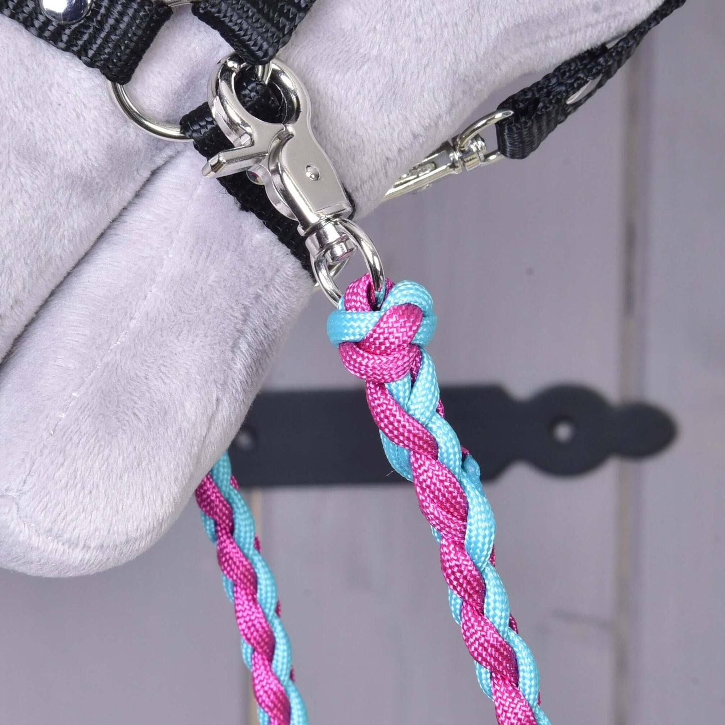 Detail Hobby Horse LarDen „Otěže knitted pinkie blue“ s kovovou karabinou připevňující polypropylenová lanová otěž k černému popruhu na světle šedém povrchu. Ideální pro sestavy otěží pro Hobby Horse. V pozadí rozmazané dřevěné dveře s černým pantem.