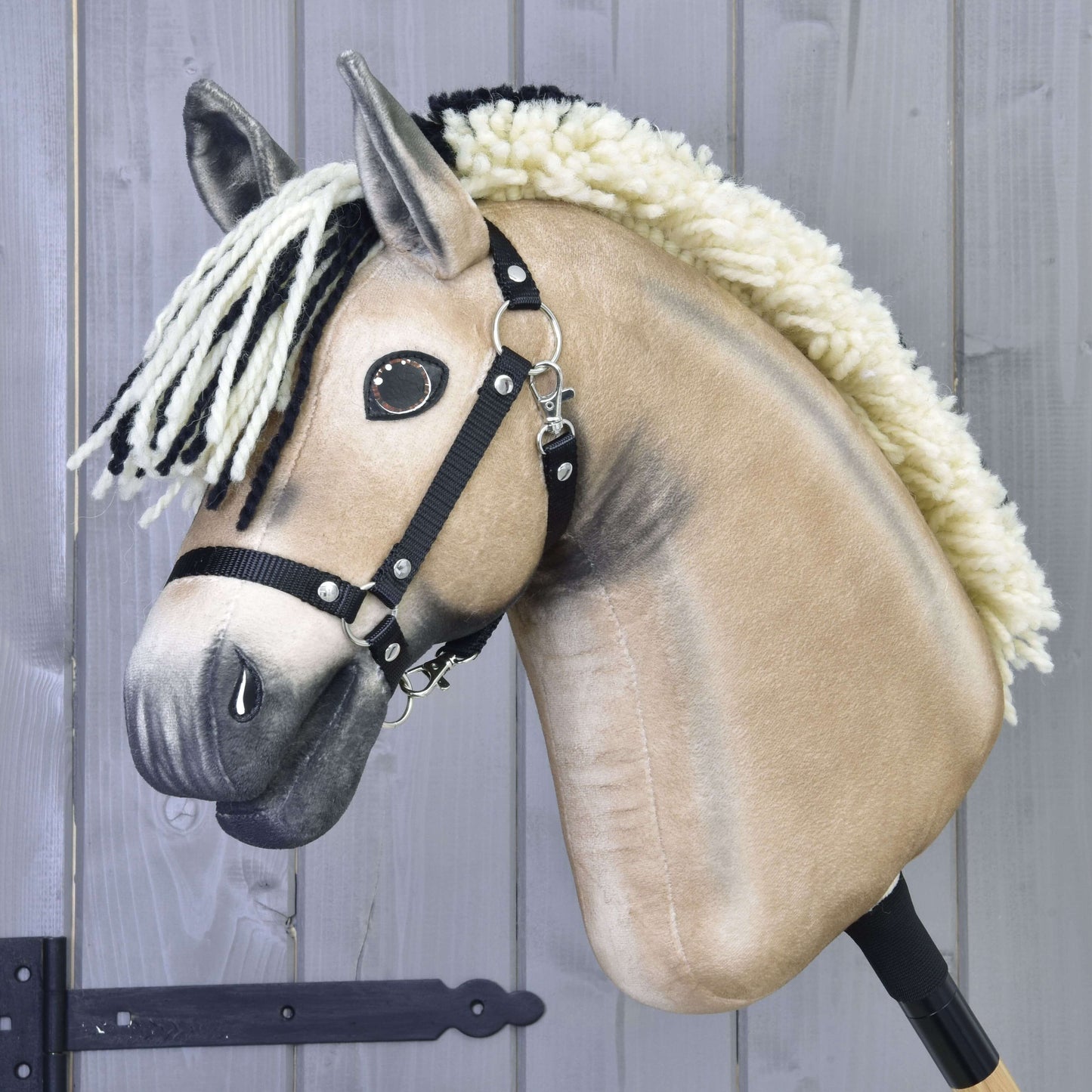 Detailní záběr na béžového Hobby Horse Ozzy s černou ohlávkou od Hobby Horse LarDen s černo-bílou přízovou hřívou, knoflíkovýma očima a tyčí, na pozadí šedého dřeva.
