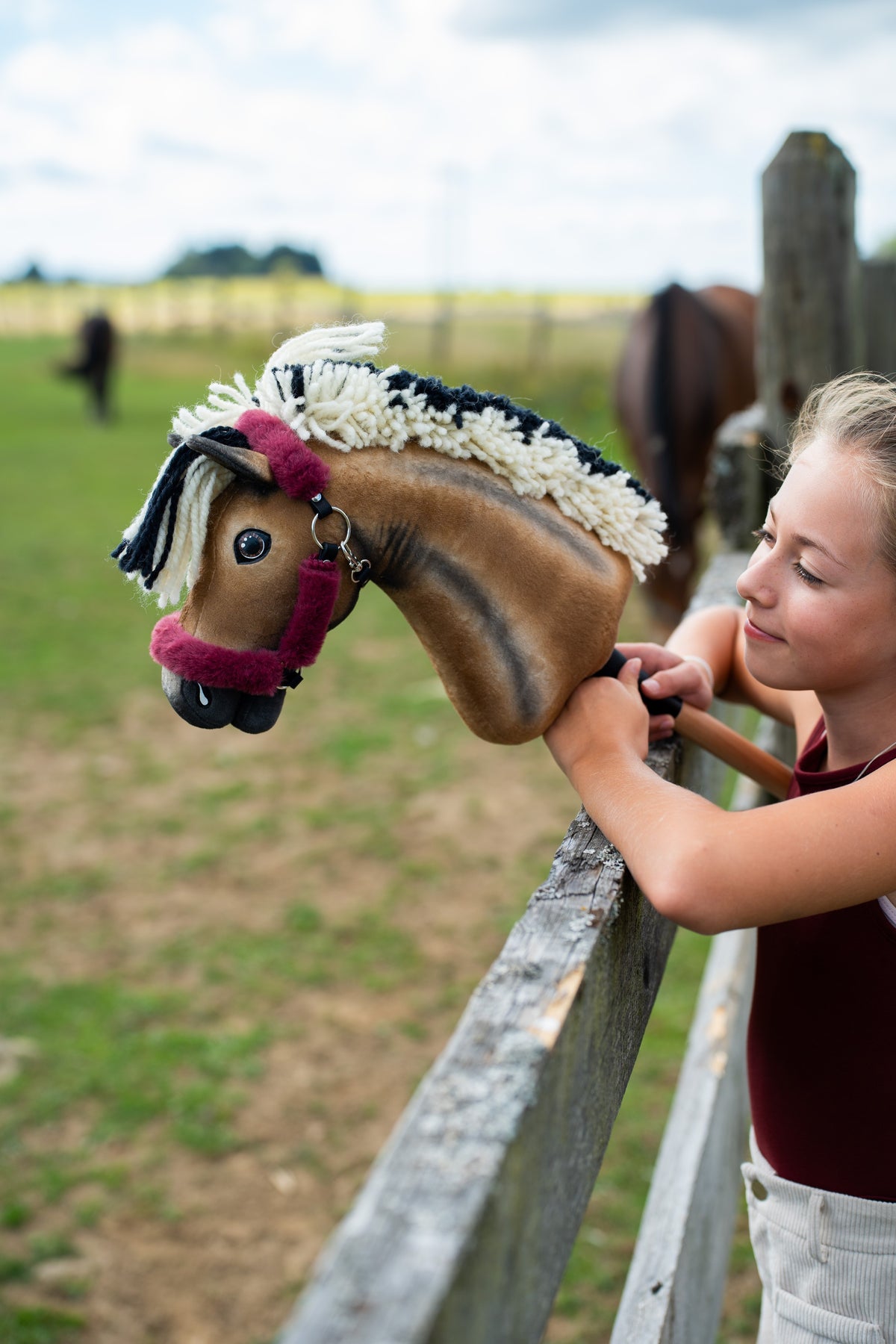 Detail photo: the head of the Hobby Horse Ozzy with Black Halter by Hobby Horse LarDen resting on a wooden paddock fence, a smiling girl and grazing horses behind - bay plush with dark shading, cream and black yarn mane - shown here with the fur-lined Teddy Maroon halter.