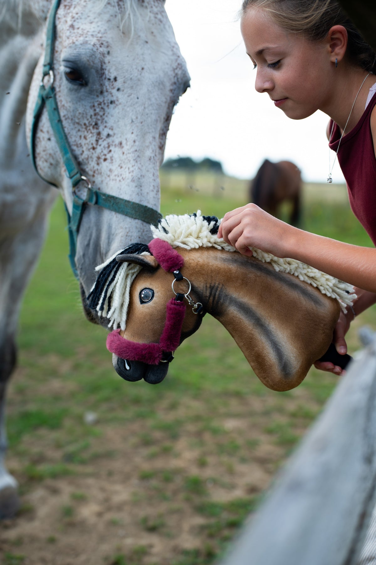 Lifestyle photo: a real grey horse lowers its head to meet the Hobby Horse Ozzy with Black Halter by Hobby Horse LarDen, held by a girl in a maroon top - bay plush head with dark shading, cream and black yarn mane - shown here with the fur-lined Teddy Maroon halter.