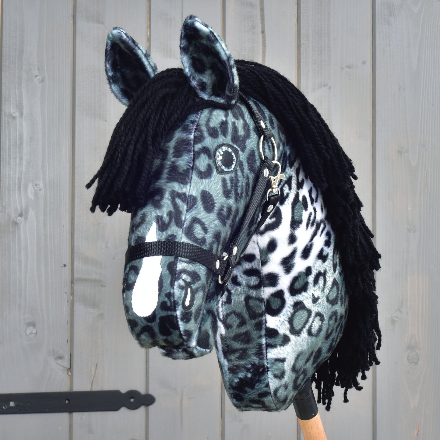 Hobby Horse Picasso s černou ohlávkou od Hobby Horse LarDen, plyšový Hobby Horse navržený tak, aby připomínal hřebce appaloosy, s výrazným černobílým leopardím vzorem, který odkazuje na vzhled skvrnité srsti. Má černou přízovou hřívu, černou ohlávku a je upevněný na dřevěné tyči, na pozadí dřevěných panelů.
