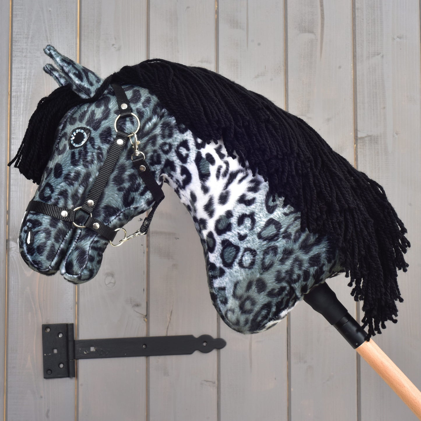 Hobby Horse Picasso s černou ohlávkou od Hobby Horse LarDen s plyšovou hlavou se šedočernou skvrnitou srstí inspirovanou appaloosou, černou ohlávkou a černou přízovou hřívou, upevněný na dřevěné tyči a zobrazený na pozadí šedé dřevěné stěny.