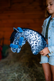 Dívka v džínové bundě drží Hobby Horse Picasso s černou ohlávkou od LarDen u červené dřevěné stěny stodoly, s modrou a bílou plyšovou hlavou s leopardím vzorem a černou přízovou hřívou, s ohlávkou Teddy Petrol podšitou kožíškem