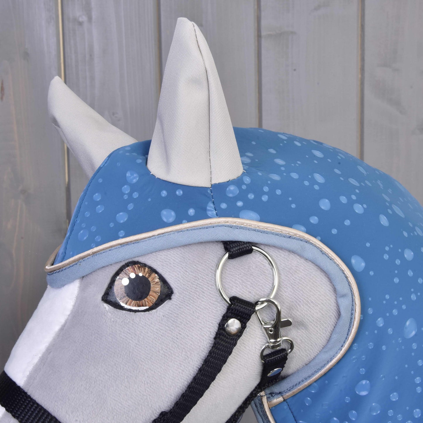 Detail hlavy Hobby Horse LarDen Pláštěnka Dolly (softshell) s modrou pláštěnkou se vzorem kapek vody a černou ohlávkou, na pozadí světlých dřevěných panelů – ideální pro hru za každého počasí.