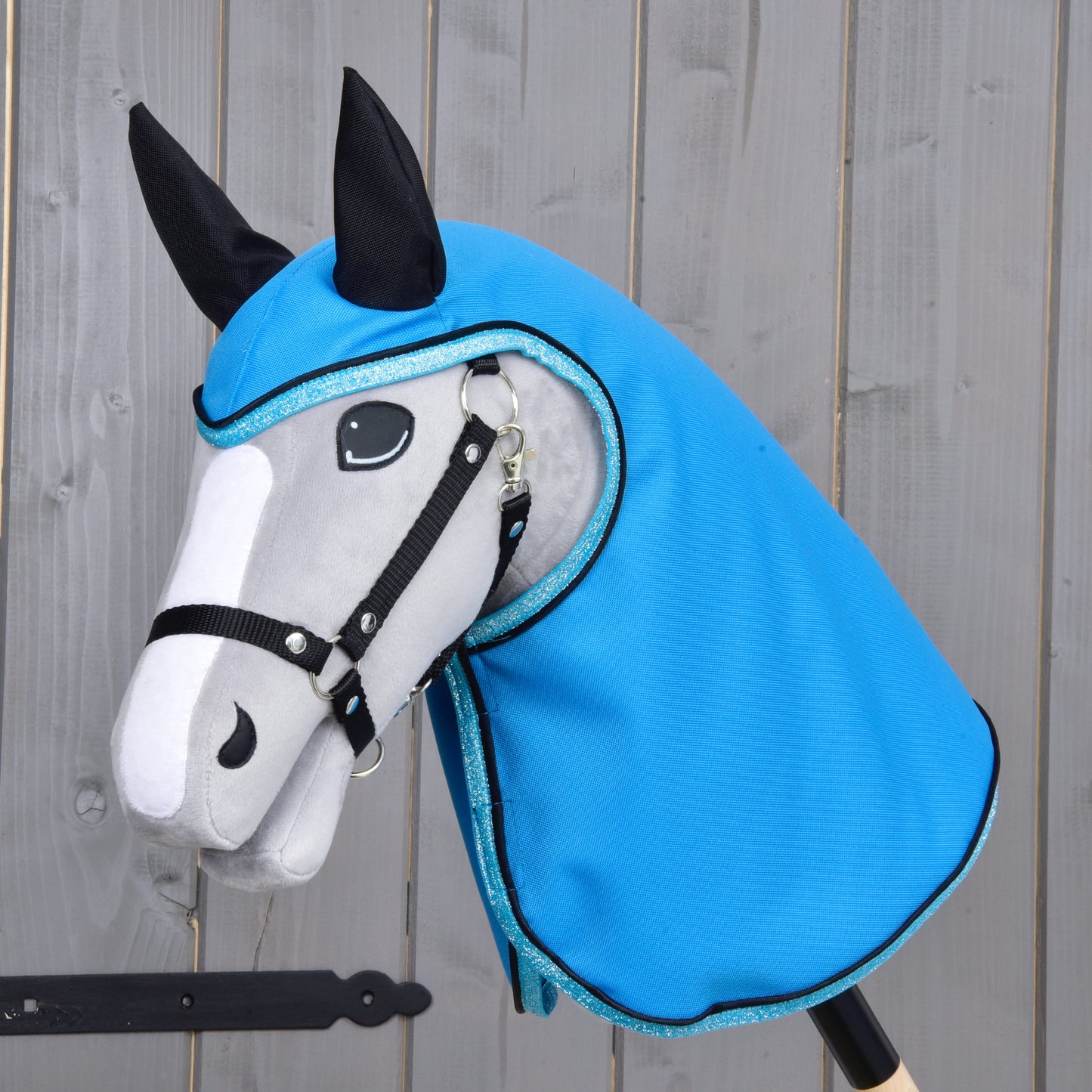 Pláštěnka Sky Blue od Hobby Horse LarDen na šedo-bílém Hobby Horse s černými otěžemi a ušima, který má na sobě lesklou kapuci ve stylu pláštěnky v nebesky modré barvě, na dřevěném pozadí.