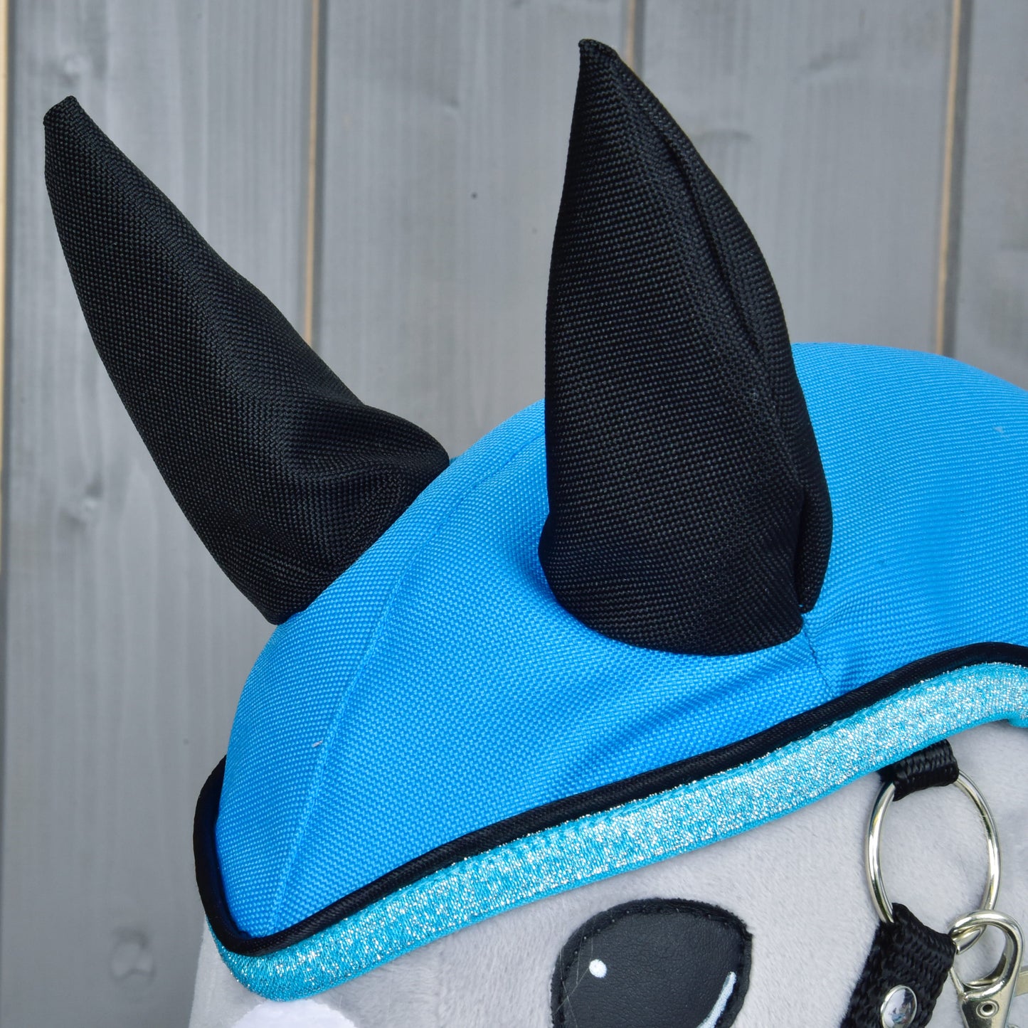 Detail plyšové hračky Hobby Horse LarDen v pláštěnce Sky Blue s černými špičatými ušima a třpytivým modrým lemováním. Pláštěnka do každého počasí vyniká na pozadí ze světle šedých dřevěných panelů.