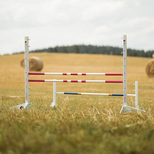 Bílé skokové tyče s červenými pruhy postavené na travnatém poli, ideální pro Hobby Horsing, s balíky sena a stromy viditelnými v pozadí pod zataženou oblohou.
