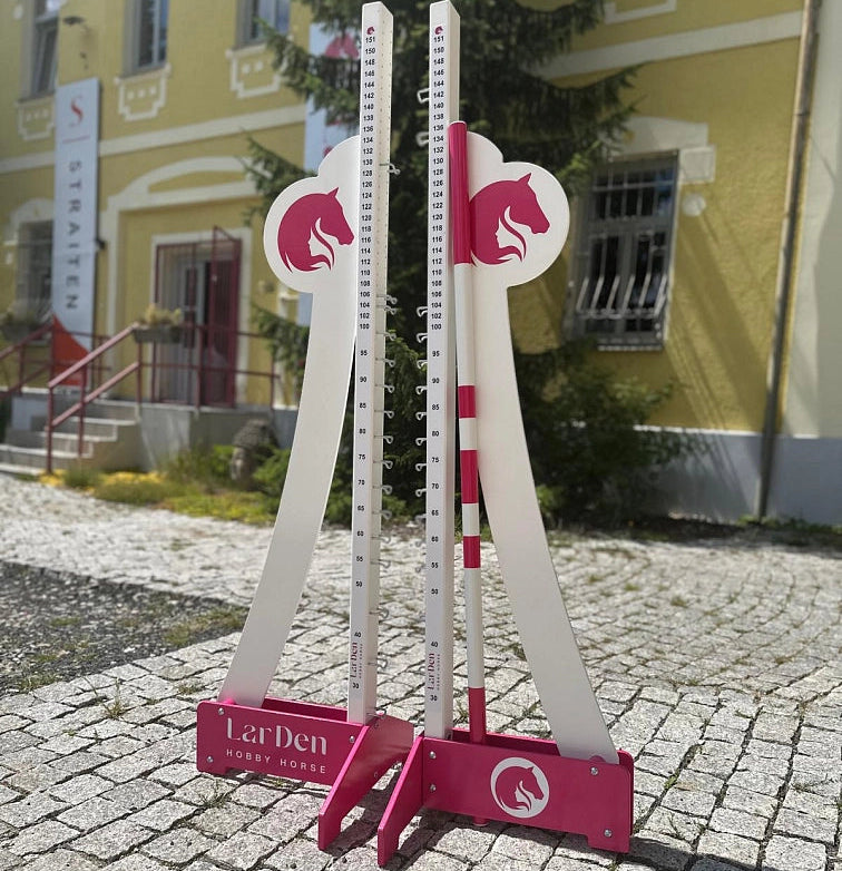 Dva bílé a růžové nastavitelné stojany na skoky pro Hobby Horsing jsou postavené venku na dlážděné cestě před žlutou budovou se schody a cedulí v pozadí.