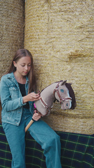 Lifestyle video: a girl in a denim jacket sits on straw bales with the Hobby Horse Guccini by Hobby Horse LarDen - grey Holsteiner-style head, white nose stripe, long dark brown yarn mane, wooden stick - shown here with the fur-lined Walnut halter, the Walnut breastgirth with fur and the Vivaldi lead rope.