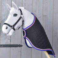 Šedá hračka Hobby Horse s černou uzdečkou, s převozní dekou Luxury od Hobby Horse LarDen — černou prošívanou, lemovanou červenou, bílou a modrou — pózuje před dřevěnou stěnou.