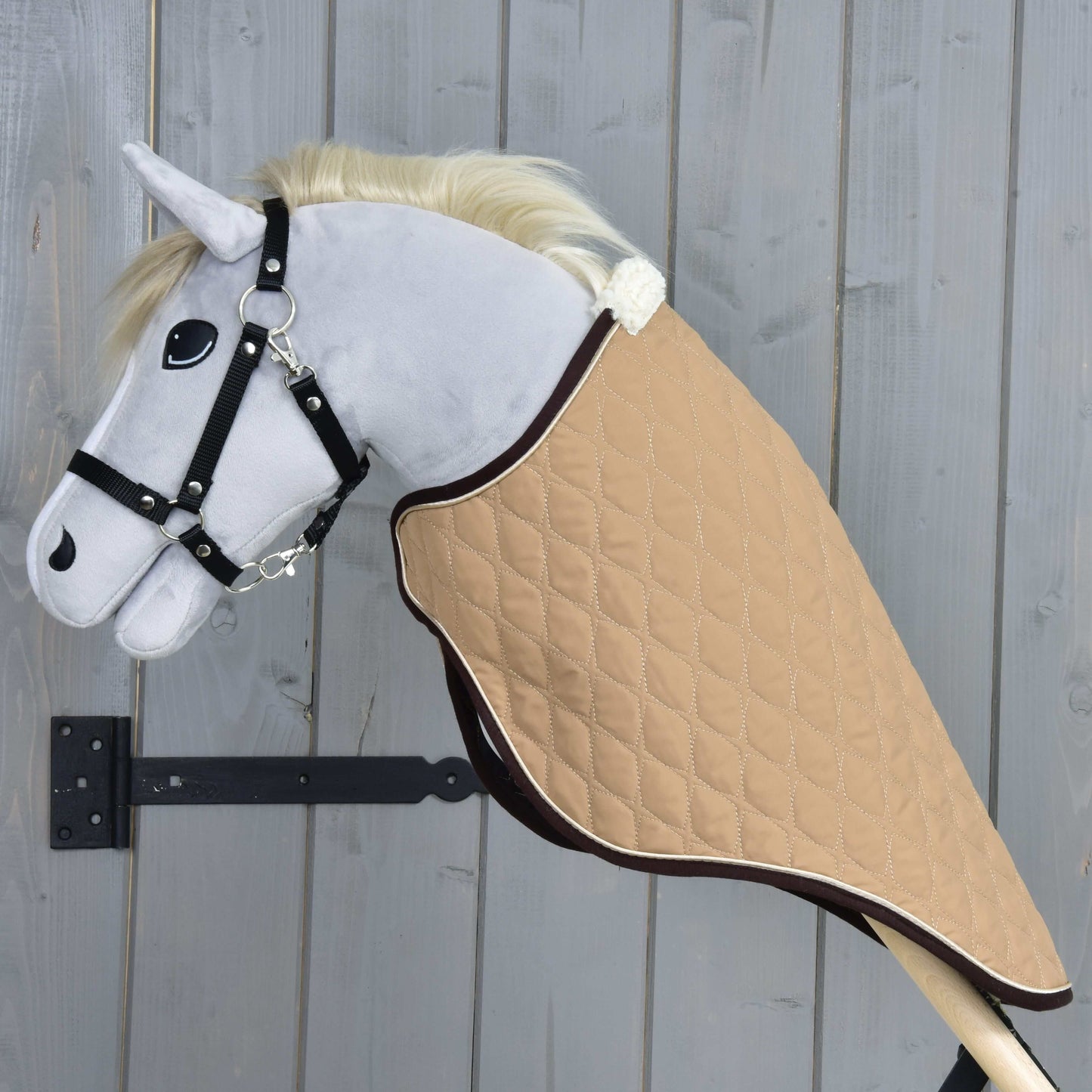 Plyšový Hobby Horse se světle šedou hlavou, bílou hřívou a černou uzdečkou má na sobě převozní deku Paris od Hobby Horse LarDen — lehkou odolnou pískovou prošívanou deku — je na dřevěné tyči a na pozadí šedé dřevěné stěny.