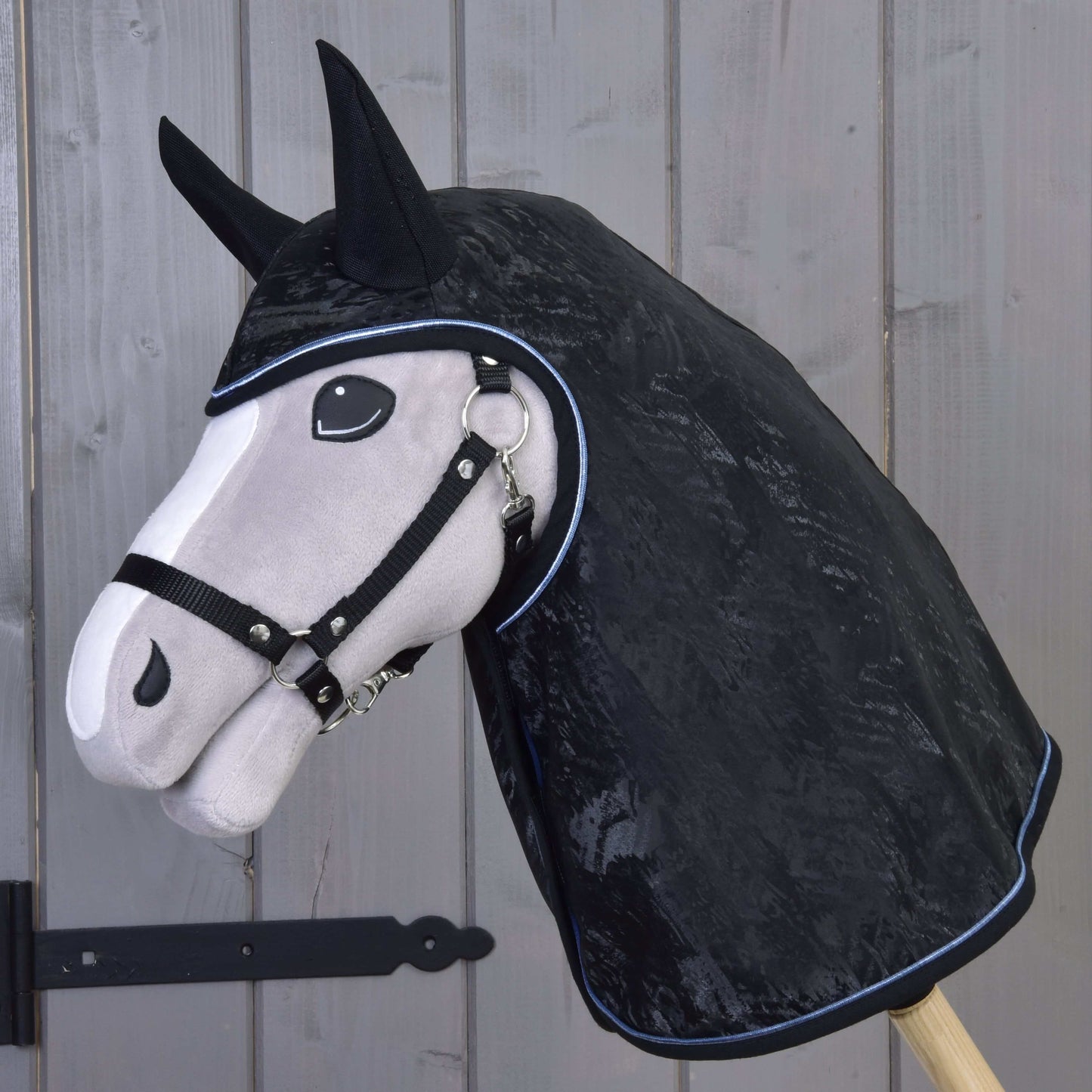 Plyšová koňská hlava na dřevěné tyči s pláštěnkou Hobby Horse LarDen Raincoat Silver Stone s kryty na uši a ohlávkou, stojící před dřevěnými panelovými dveřmi.