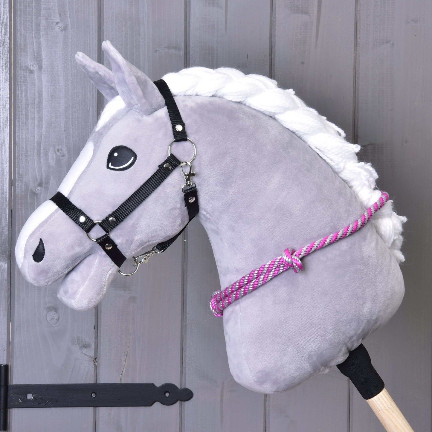 Nákrčník s růžovým proužkem od Hobby Horse LarDen na šedé plyšové koňské hlavě s bílou hřívou, černou uzdečkou a růžovo-bílým nákrčníkem na dřevěné tyči, ideální pro nácvik jízdy bez uzdečky před šedou dřevěnou stěnou.