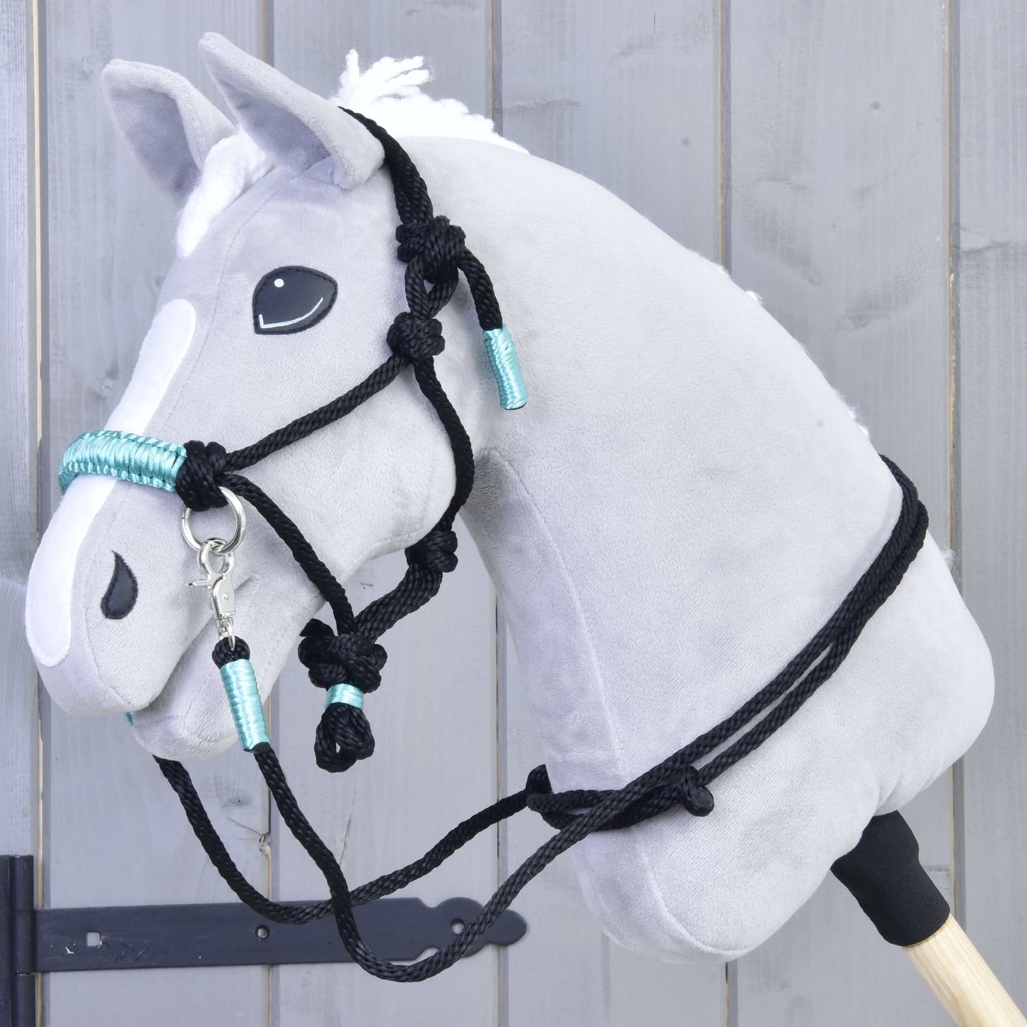 Provazový set Turquoise od Hobby Horse LarDen je zobrazen na šedém plyšovém Hobby Horse s bílou hřívou, černou uzdečkou a tyrkysovými detaily, upevněném na dřevěné tyči před šedou dřevěnou stěnou.