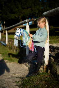 Lifestyle photo: a girl in a mint sweatshirt stands at a white rail by the riding arena holding the Hobby Horse Rainbow New by Hobby Horse LarDen - white plush head with a long pastel rainbow yarn mane - shown here with the Halter blue with fur and the Lead rope stripe blue.