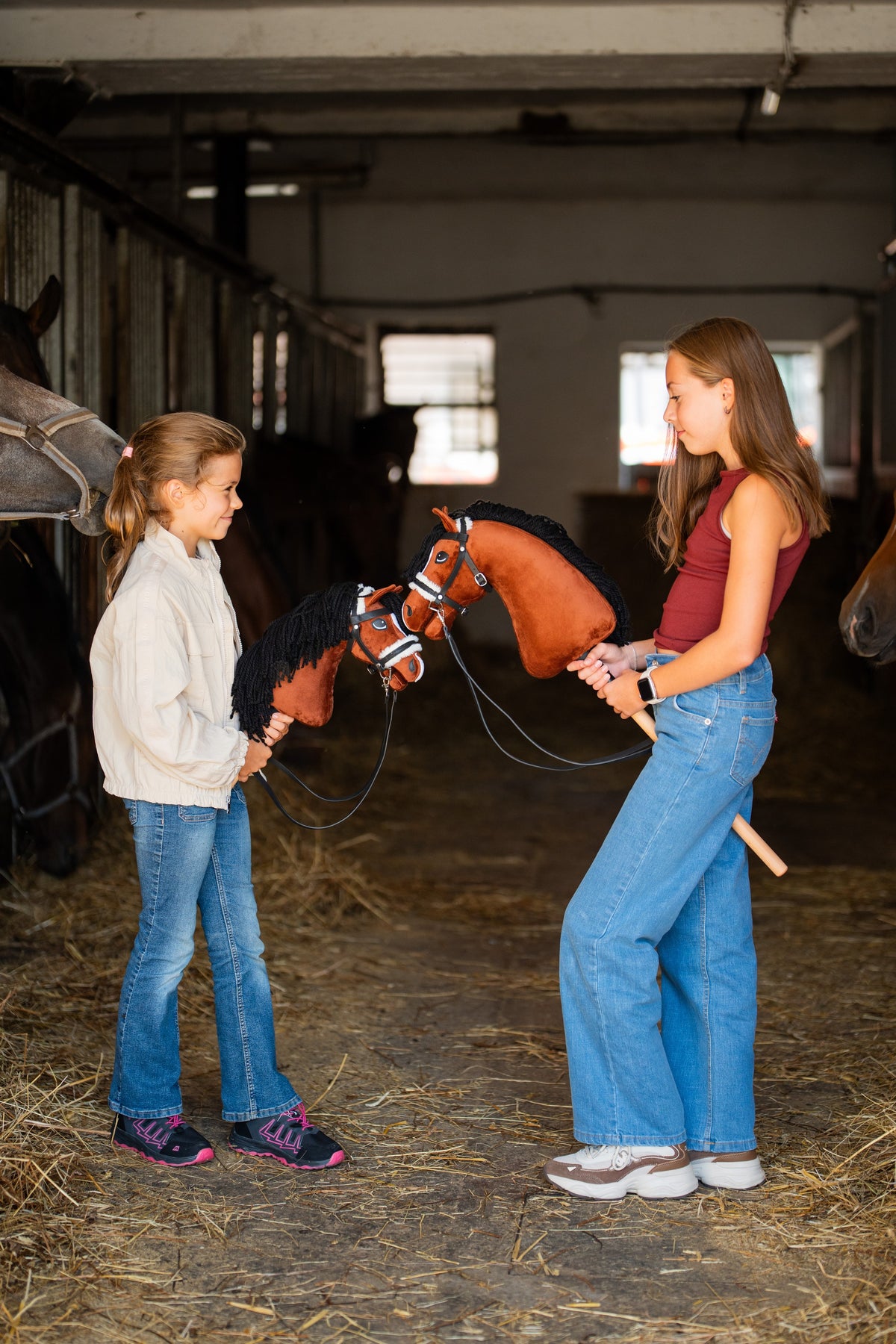 Dvě dívky stojí proti sobě v uličce stáje a každá drží Hobby Horse Rocky od Hobby Horse LarDen, s ryzákově hnědou plyšovou hlavou s bílou lysinou a černou přízovou hřívou, s kompletní černou uzdečkou s kožešinou a otěžemi