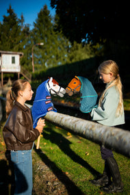 Dvě dívky u dřevěného hrazení drží v večerním slunci Hobby Horse Rufus s černou Ohlávkou v dece s odnímatelným krytem krku Bluebell a Hobby Horse Pippa s černou Ohlávkou v dece s odnímatelným krytem krku Lagoon čumákem k sobě, oba od Hobby Horse LarDen