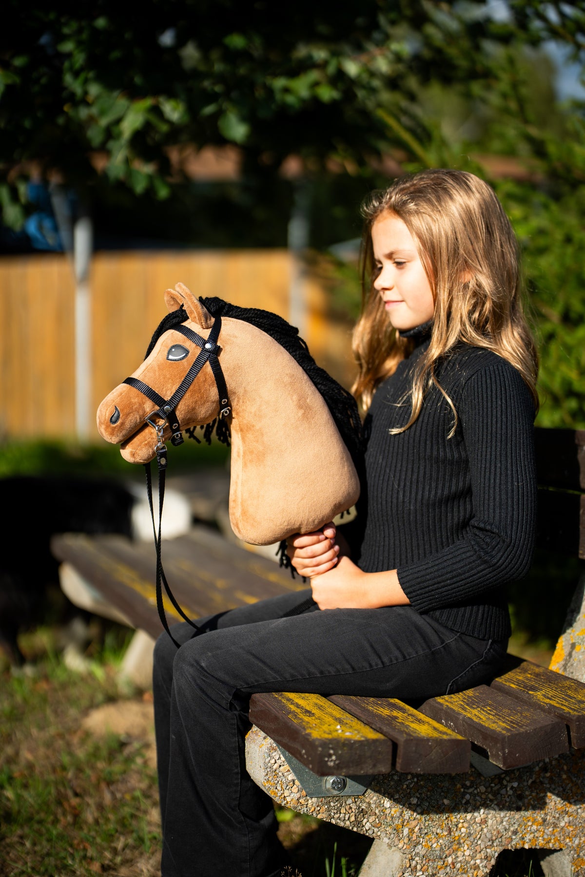 Lifestyle photo: a girl in a black jumper sits on a park bench in the evening sun holding the Hobby Horse Samantha with Black Halter by Hobby Horse LarDen - sandy plush head, black yarn mane - shown here with the black bridle with strap reins.