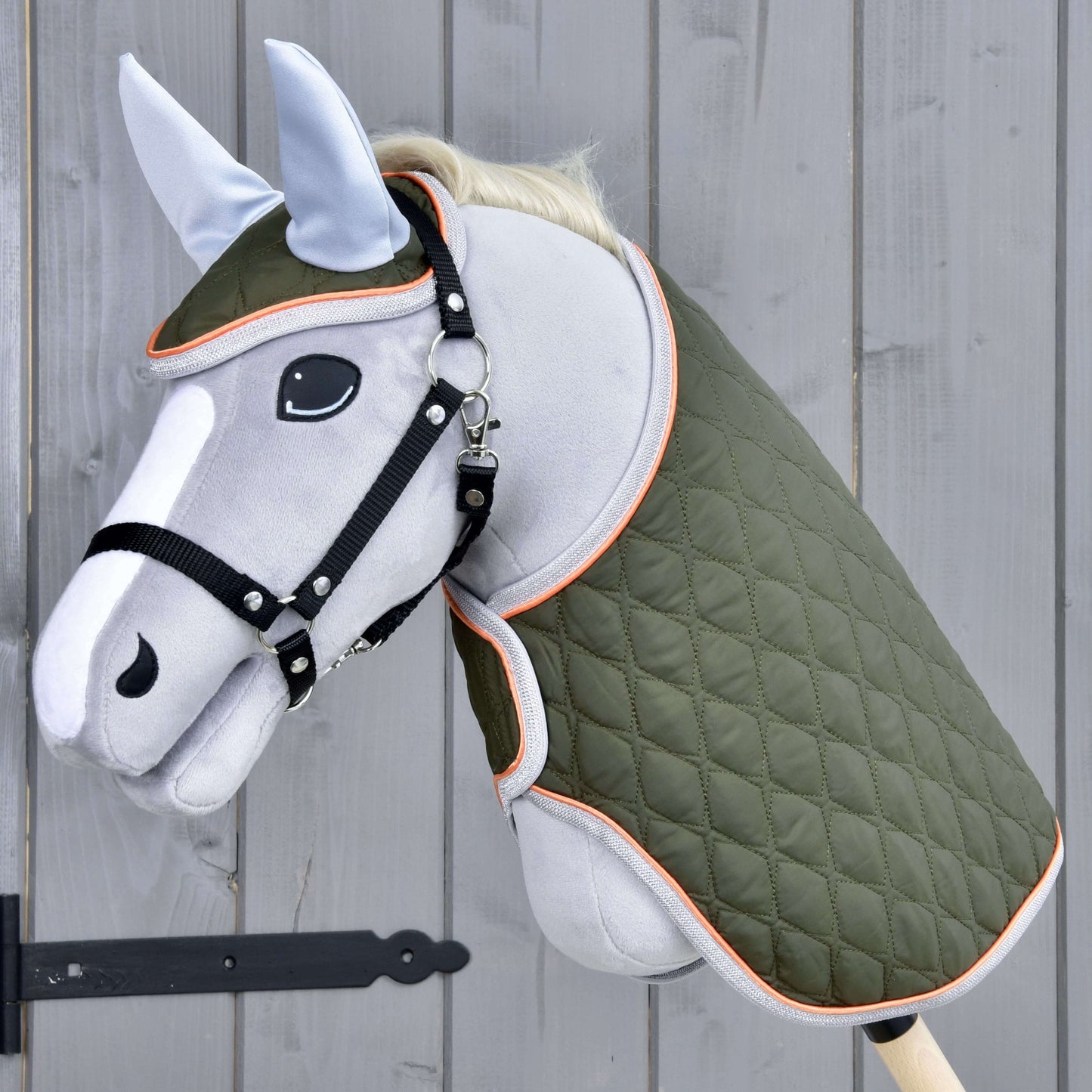 Set Moos od Hobby Horse LarDen s šedým Hobby Horse na dřevěné tyči, černou uzdečkou, zelenou prošívanou dekou a světle modrou čabrakou na šedé dřevěné stěně.