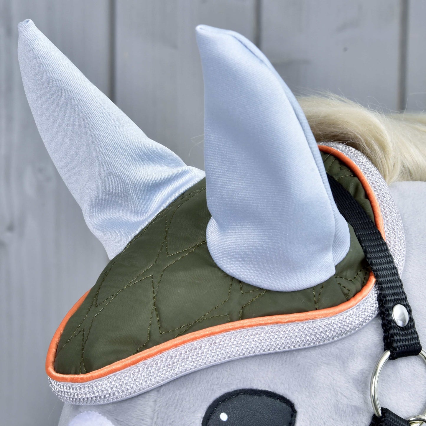Set Moos od Hobby Horse LarDen, detail hlavy plyšového koně s olivově zelenou a oranžovou čabrakou, dvěma světle šedými špičatými látkovými ušima a roztomilou koňskou dekorací na světle šedém dřevěném pozadí.