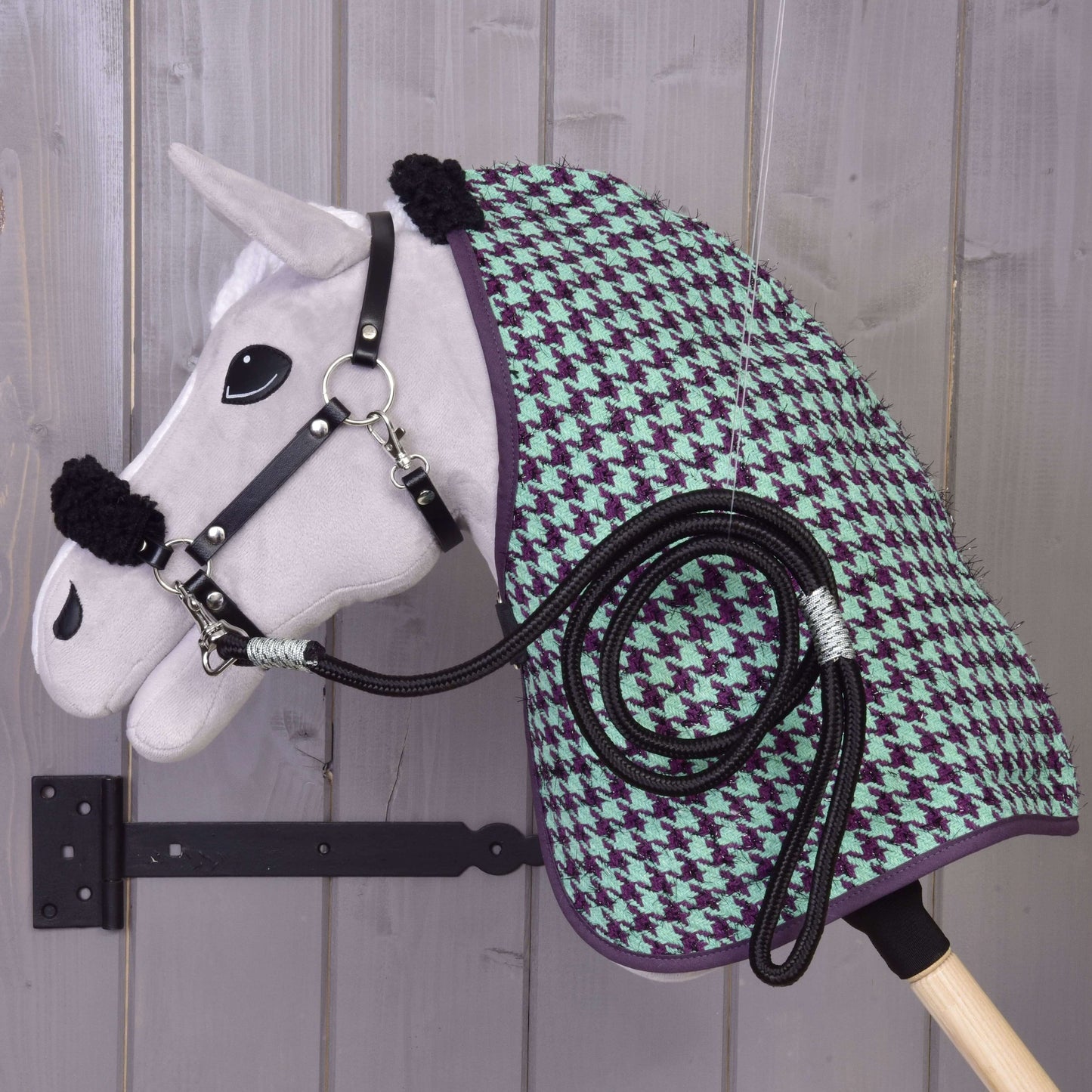 Set Harmony Green od Hobby Horse LarDen – plyšový Hobby Horse s černou ohlávkou, zeleno-fialovou stájovou dekou se vzorem kohoutí stopy a stočeným černým vodítkem – je zavěšený na dřevěném nástěnném držáku před šedou dřevěnou stěnou.