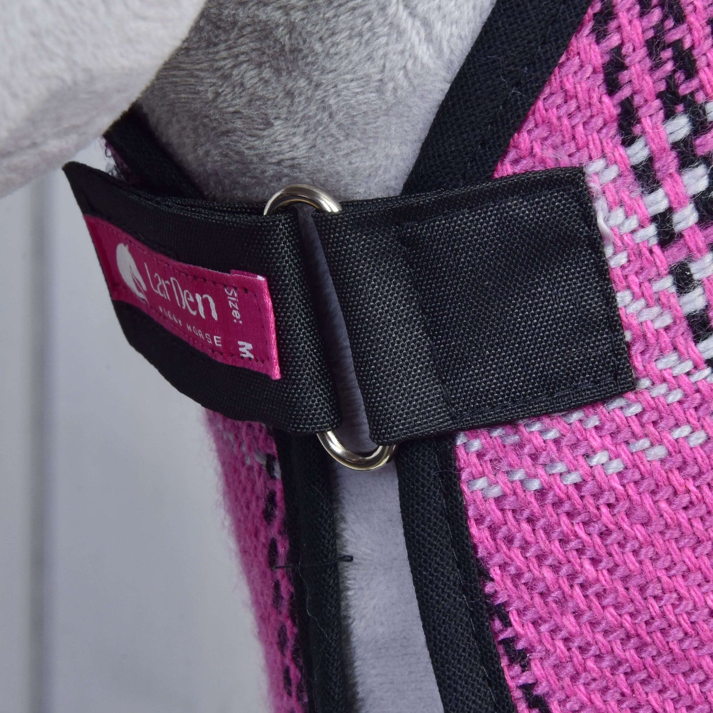 Detail Set Harmony Pink od Hobby Horse LarDen: růžová tkaná látka s černým řemínkem ohlávky, kovovým kroužkem a růžovým štítkem. Pozadí naznačuje šedé a černé detaily oděvu nebo doplňku pro Hobby Horse, pravděpodobně stájové deky.