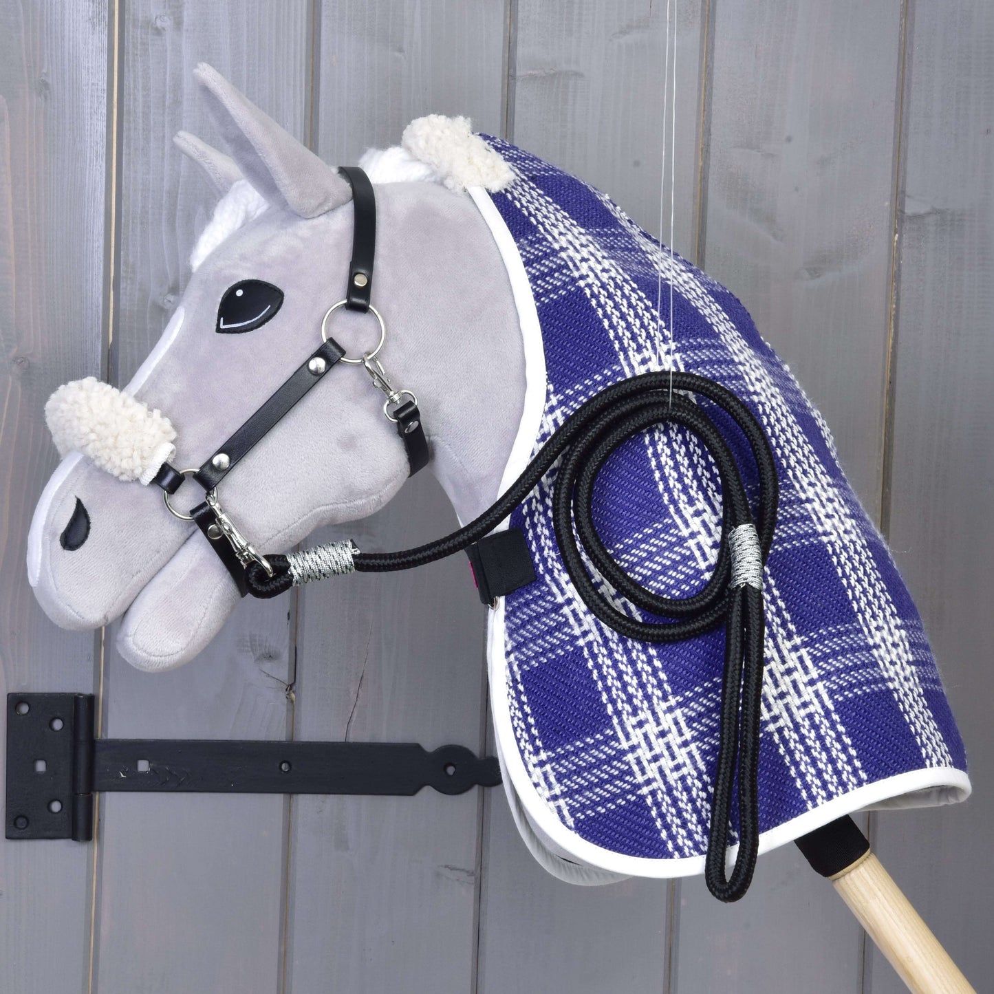 Set Harmony Victory od Hobby Horse LarDen tvoří šedý Hobby Horse s černou ohlávkou a otěžemi, modro-bílou kostkovanou stájovou dekou a bílou umělou kožešinou na uších a krku, zavěšený na stěně na pozadí šedého dřevěného obložení.