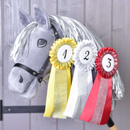 Šedý plyšový Hobby Horse s bílou přízovou hřívou má na krku zavěšený Hobby Horse LarDen Set Hobby Horse Kokarda NEW – žlutou, bílou a červenou kokardu pro první, druhé a třetí místo – na dřevěném pozadí.