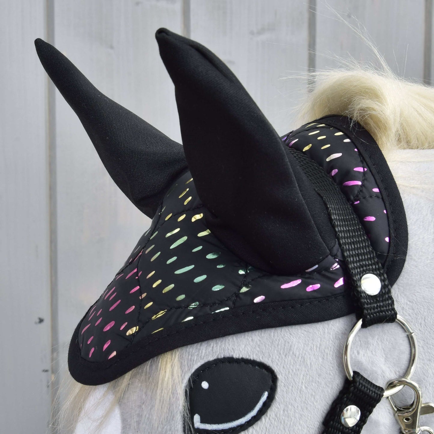 Detail Hobby Horse LarDen „Set Dreamy“ na Hobby Horse s bílým tělem, černou čabrakou s barevnými reflexními puntíky, usměvavým plstěným okem a uzdečkou, před světlým dřevěným obkladem.