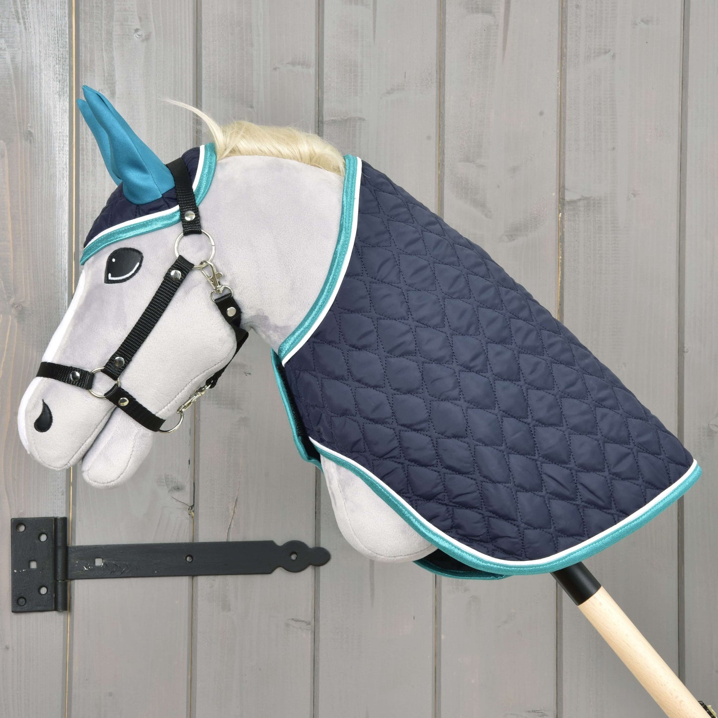 Set Supreme Turquoise od Hobby Horse LarDen s šedým plyšovým Hobby Horse s modrýma ušima, černou uzdečkou a prošívanou tmavě modrou dekou s tyrkysovým lemem, nasazený na dřevěné tyči před stěnou ze světlého dřeva.