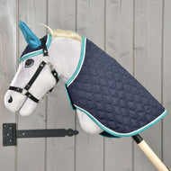 Set Supreme Turquoise od Hobby Horse LarDen s šedým plyšovým Hobby Horse s modrýma ušima, černou uzdečkou a prošívanou tmavě modrou dekou s tyrkysovým lemem, nasazený na dřevěné tyči před stěnou ze světlého dřeva.