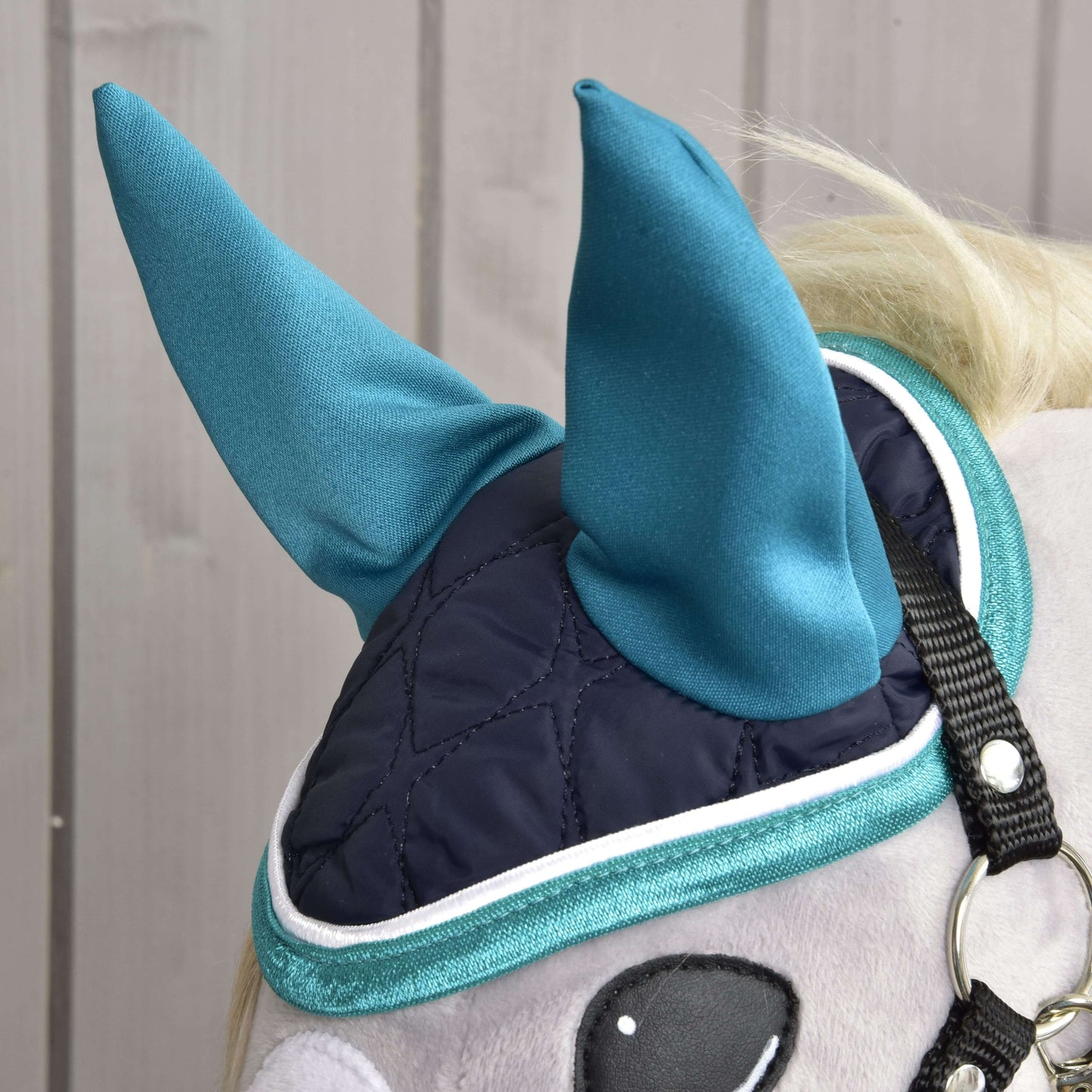 Detail Set Supreme Turquoise od Hobby Horse LarDen: plyšový koník má tmavě modrou prošívanou masku na oči s tyrkysovými špičatými kryty uší a lemem – stylovou sadu čabraky – pro zábavu a ochranu, na pozadí světlého dřeva.