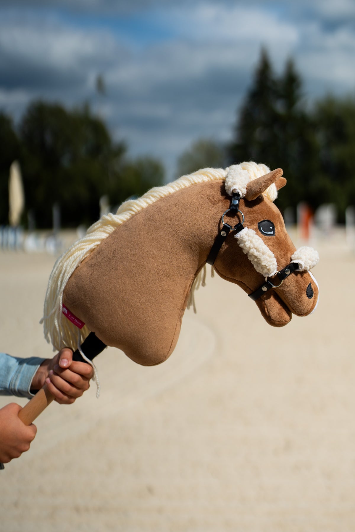 Hlava Hobby Horse Smart s černou ohlávkou od LarDen je zvednutá na pískové jízdárně pod tmavými mraky, s palomino plyšem, krémovou přízovou hřívou a bílým kožešinovým nánosníkem, s černou koženkovou ohlávkou s celoplošným kožíškem