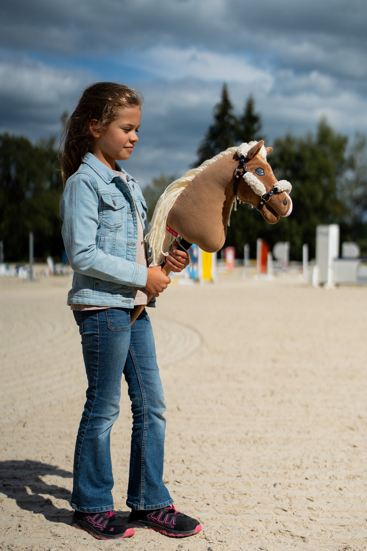 Dívka v džínové bundě stojí na pískové jízdárně a drží Hobby Horse Smart s černou ohlávkou od LarDen, za ní jsou skokové tyče, s palomino plyšovou hlavou a dlouhou krémovou přízovou hřívou, s černou koženkovou ohlávkou s celoplošným kožíškem
