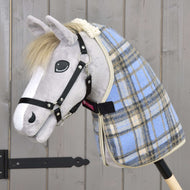 Plyšový Hobby Horse se šedým obličejem, bílou hřívou a černou uzdečkou má na sobě Hobby Horse LarDen Deka stájová Light Blue, je nasazený na dřevěné tyči a stojí před rustikální dřevěnou stěnou.