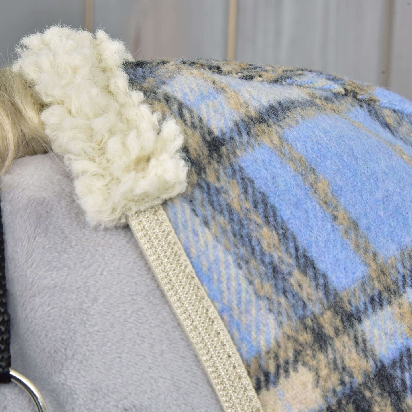 Detail deky Hobby Horse LarDen Deka stájová Light Blue s modrým, béžovým a šedým kostkovaným vzorem a krémovým sherpa lemem, přehozené přes šedý povrch. Vlevo je částečně vidět černý popruh a stříbrný kroužek.