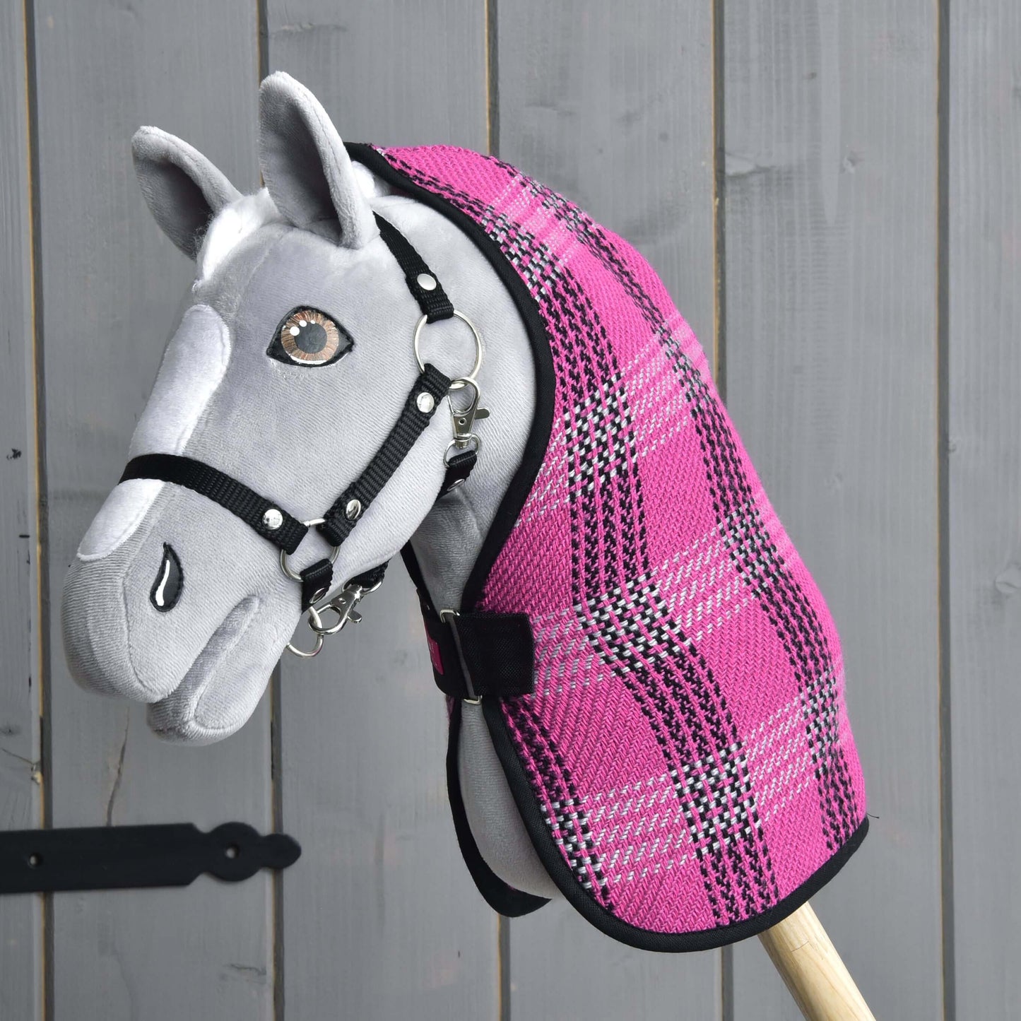Šedý Hobby Horse od Hobby Horse LarDen s černou uzdečkou a dekou Deka stájová Pink, na dřevěné tyči, před šedou dřevěnou stěnou.