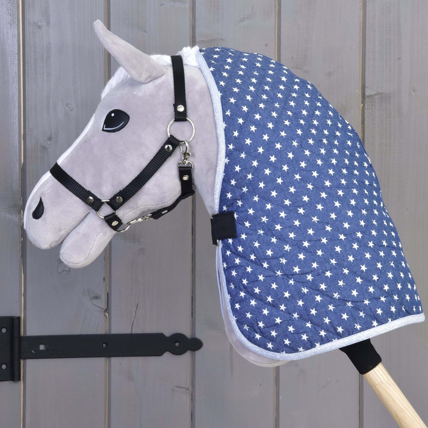 Šedá hračka Hobby Horse od Hobby Horse LarDen s černou uzdečkou a dekou Deka stájová Star, upevněná na dřevěné tyči, na pozadí šedých dřevěných dveří.