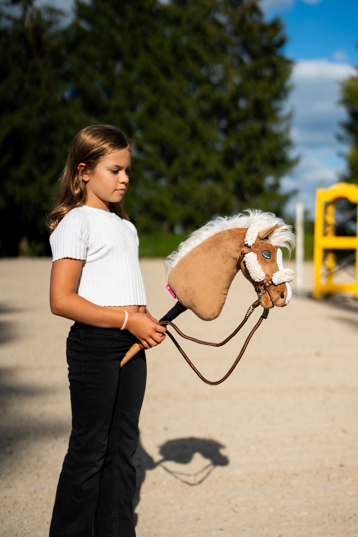 Lifestyle photo: a girl in a white top walks the Hobby Horse Star with black halter by Hobby Horse LarDen across a sand arena, a yellow jump wing behind - palomino plush head, flowing white yarn mane - shown here with the brown halter with complete fur and the Vivaldi reins.